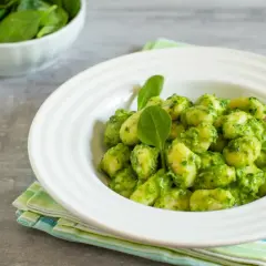Puszyste gnocchi z pesto, podane w białej misce, ozdobione świeżymi listkami szpinaku. Obok miseczka ze świeżym szpinakiem.