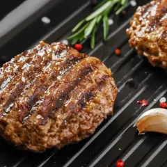 Soczyste mięso na burgery grilluje się na patelni, obok gałązka rozmarynu i ząbek czosnku.
