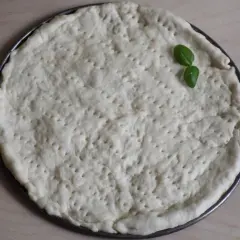 Surowy placek na pizzę w okrągłej formie, z kilkoma listkami bazylii. Proste ciasto na pizzę zawsze wychodzi.