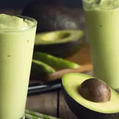Dwa szklanki kremowego smoothie z awokado, idealna przekąska na upalne dni. Obok leżą świeże awokado i zielone słomki.