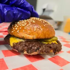 Rękawiczka w niebieskiej rękawiczce dociska pysznego smash burgera z serem i ogórkiem. Oto jak wygląda smash burger co to!
