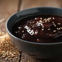 Gęsty, błyszczący sos teriyaki w misce, posypany sezamem. Obok garść prażonych ziaren sezamu. Idealny do dań kuchni azjatyckiej.