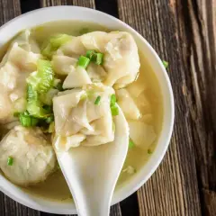 Aromatyczna zupa wonton z zieloną cebulką i kawałkami selera, podana w białej miseczce na drewnianym blacie.