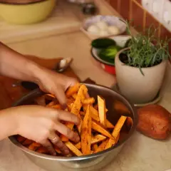 Ręce mieszają pokrojone bataty w misce, przygotowując je do pieczenia. Obok leży cały batat, a w tle zioła i warzywa, jakby czekały na sos belgijski.