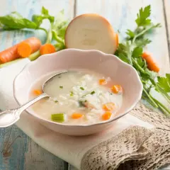 Aromaticzna zupa na rosole z ryżem, marchewką i selerem. Obok miseczki świeże warzywa: marchew, cebula, natka pietruszki.
