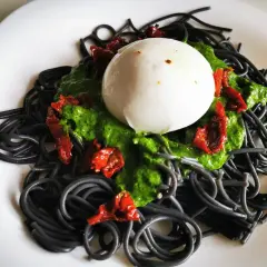 Czarny makaron z zielonym pesto, suszonymi pomidorami i kulką mozzarelli na białym talerzu.