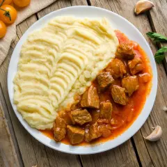 Pyszny gulasz z karkówki z kremowym puree ziemniaczanym na białym talerzu, ozdobiony pomidorkami i czosnkiem.