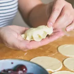 Dłonie formują pieroga z ciasta na pierogi bez jajka. Na blacie leżą okrągłe placki z ciasta i miska z wiśniami.