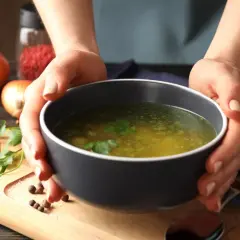 Dłonie trzymają miskę z aromatycznym rosołem, obok cebula i przyprawy. Idealny przepis na to, jak ugotować rosół.