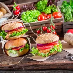 Dwa apetyczne burgery z sałatą, pomidorem i cebulą na drewnianej desce, w tle warzywa.