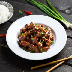Pyszny kurczak luc lac w sosie, posypany sezamem i szczypiorkiem, obok miseczka ryżu, papryczki chili i pałeczki.
