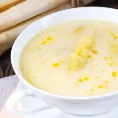 Aksamitna, tradycyjna zupa szparagowa z kawałkami szparagów i kropelkami oliwy, obok pęczka świeżych białych szparagów.