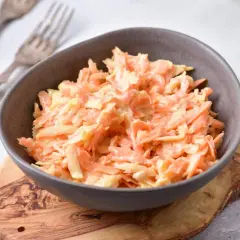 Pyszna surówka z marchewki z majonezem, podana w misce na drewnianej desce. Obok widoczne widelce i zielone jabłko.