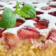 Wilgotne ciasto jogurtowe z budyniem i truskawkami, posypane cukrem pudrem i ozdobione miętą.