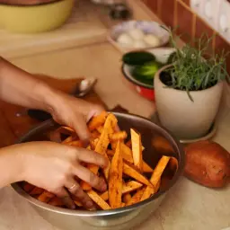 Ręce mieszają pokrojone bataty w misce, przygotowując je do pieczenia. Obok leży cały batat, a w tle zioła i warzywa, jakby czekały na sos belgijski.