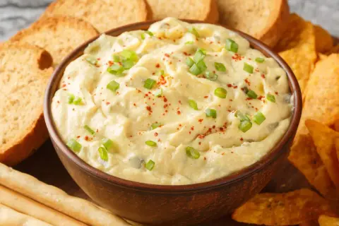 Kremowy dip z zieloną cebulką i papryką, otoczony chrupiącymi grzankami, nachos i paluszkami. Idealny do zanurzania!