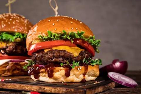Dwa soczyste burgery z serem, sałatą i pomidorem na desce, obok cebuli i papryczki chili.