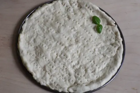 Surowy placek na pizzę w okrągłej formie, z kilkoma listkami bazylii. Proste ciasto na pizzę zawsze wychodzi.