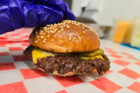 Rękawiczka w niebieskiej rękawiczce dociska pysznego smash burgera z serem i ogórkiem. Oto jak wygląda smash burger co to!