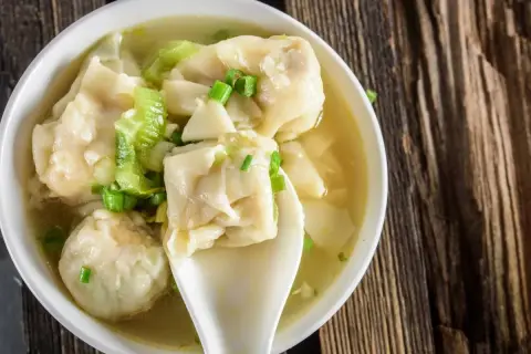 Aromatyczna zupa wonton z zieloną cebulką i kawałkami selera, podana w białej miseczce na drewnianym blacie.