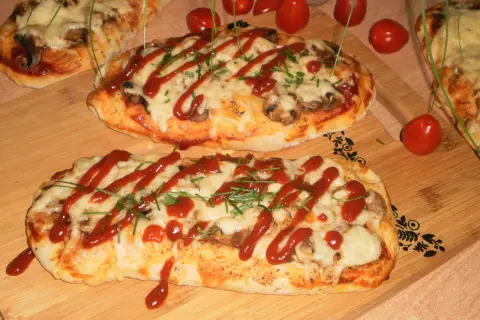 Dwa podłużne, zapieczone ciasta na pizzę z serem, pieczarkami i keczupem, ozdobione szczypiorkiem.