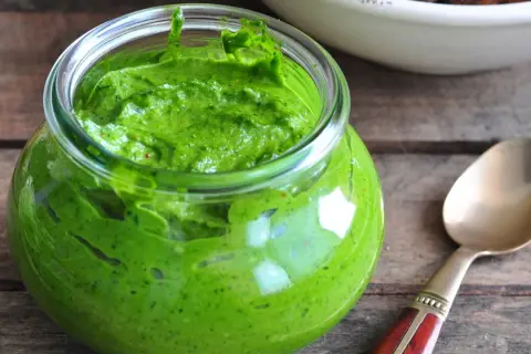 Słoik pełen intensywnie zielonego pesto z pietruszki, gotowego do użycia. Obok leży złota łyżeczka.