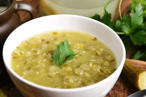 Zupa ogórkowa przepis babci. Aromatyczna, domowa zupa z kiszonych ogórków, podana w białej miseczce z gałązką natki pietruszki. Obok leżą ogórki kiszone.