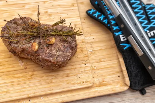 Soczysty rostbef wołowy z rozmarynem i czosnkiem na desce, obok rękawicy grillowej.