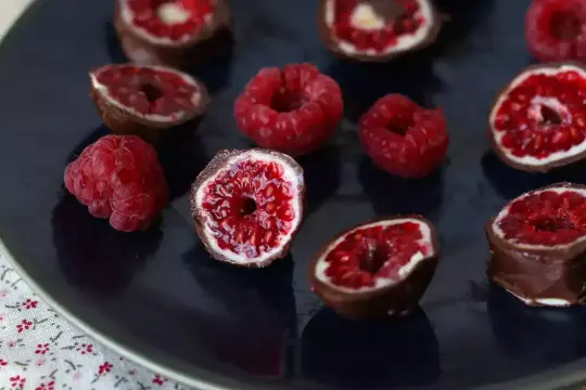 Maliny mrożone w czekoladzie, niektóre przekrojone, ukazujące soczyste wnętrze. Pyszna przekąska.