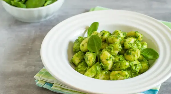 Puszyste gnocchi z pesto, podane w białej misce, ozdobione świeżymi listkami szpinaku. Obok miseczka ze świeżym szpinakiem.