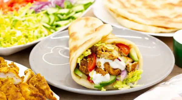 Pyszna shoarma drobiowa w picie z warzywami i sosem. Obok talerz z dodatkami: sałatka, pomidory, ogórki, cebula i placki chleba.