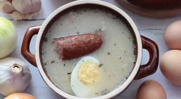 Aromaticzny żurek z kiełbasą i jajkiem, gotowy do podania. W tle czosnek, cebula i jajka, składniki idealnego zakwasu na żurek.