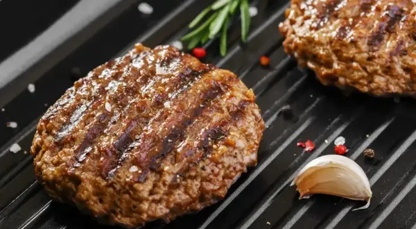 Soczyste mięso na burgery grilluje się na patelni, obok gałązka rozmarynu i ząbek czosnku.