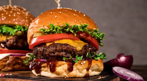 Dwa soczyste burgery z serem, sałatą i pomidorem na desce, obok cebuli i papryczki chili.