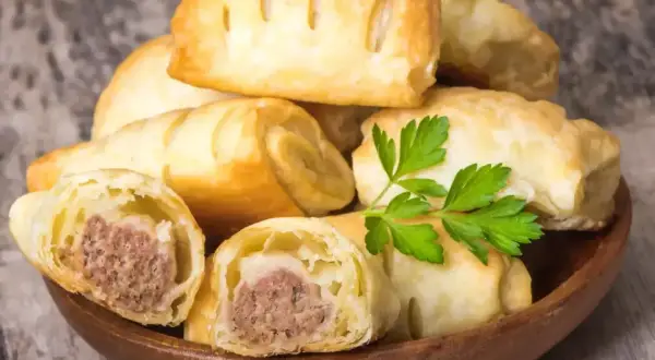 Stos pieczonego ciasta francuskiego z kiełbasą w środku, ozdobiony gałązką pietruszki.