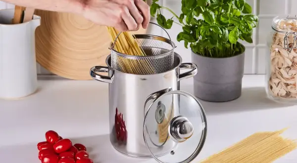 Ręka wkłada spaghetti do garnka, by ugotować je idealnie al dente. Obok pomidorki i świeża bazylia.