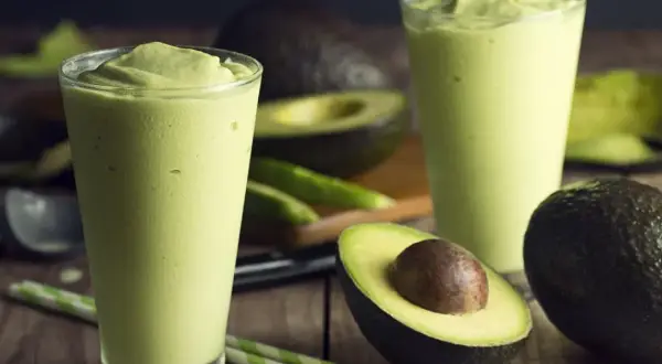 Dwa szklanki kremowego smoothie z awokado, idealna przekąska na upalne dni. Obok leżą świeże awokado i zielone słomki.