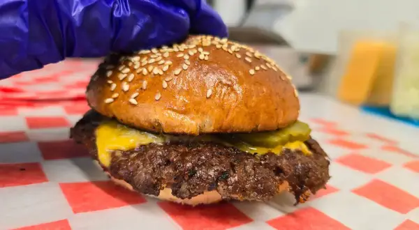 Rękawiczka w niebieskiej rękawiczce dociska pysznego smash burgera z serem i ogórkiem. Oto jak wygląda smash burger co to!