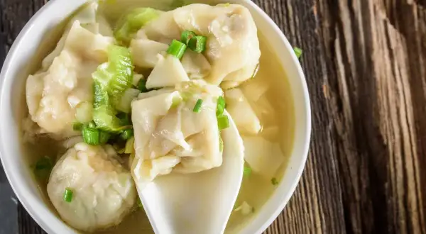Aromatyczna zupa wonton z zieloną cebulką i kawałkami selera, podana w białej miseczce na drewnianym blacie.