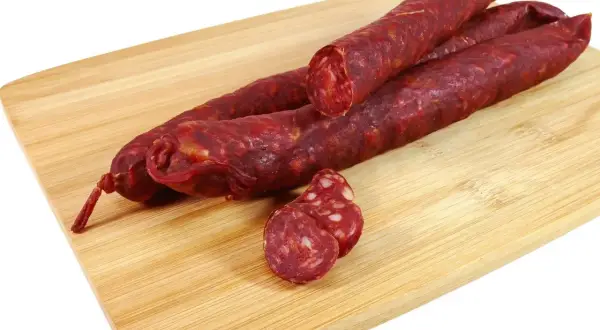 Trzy długie kiełbasy chorizo leżą na drewnianej desce. Jedna jest przekrojona, ukazując charakterystyczne kawałki tłuszczu.