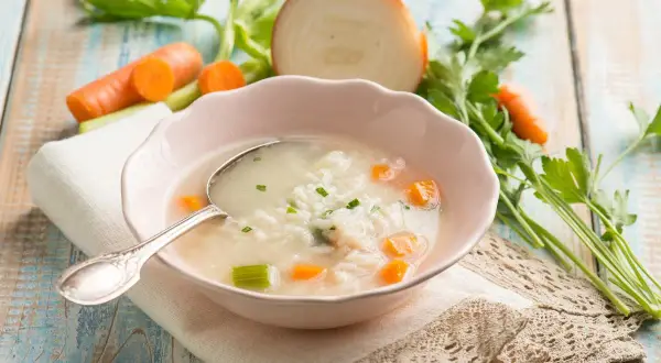 Aromaticzna zupa na rosole z ryżem, marchewką i selerem. Obok miseczki świeże warzywa: marchew, cebula, natka pietruszki.