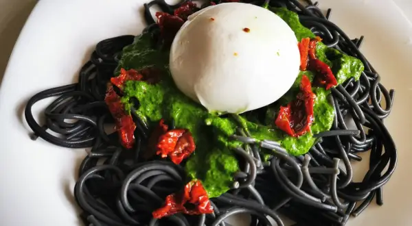 Czarny makaron z zielonym pesto, suszonymi pomidorami i kulką mozzarelli na białym talerzu.