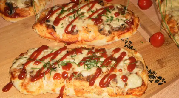 Dwa podłużne, zapieczone ciasta na pizzę z serem, pieczarkami i keczupem, ozdobione szczypiorkiem.