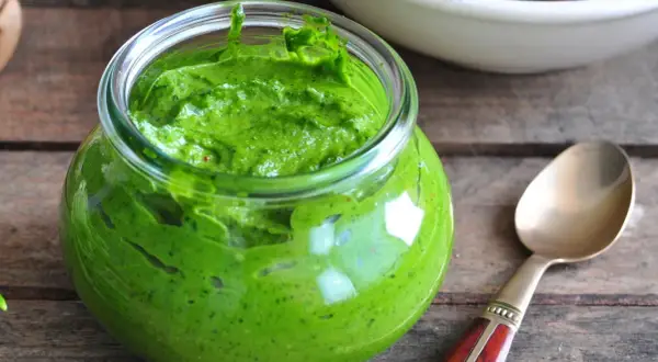 Słoik pełen intensywnie zielonego pesto z pietruszki, gotowego do użycia. Obok leży złota łyżeczka.