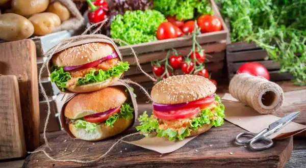 Dwa apetyczne burgery z sałatą, pomidorem i cebulą na drewnianej desce, w tle warzywa.