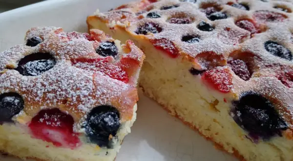 Puszyste ciasto na maślance z jagodami i malinami, posypane cukrem pudrem.