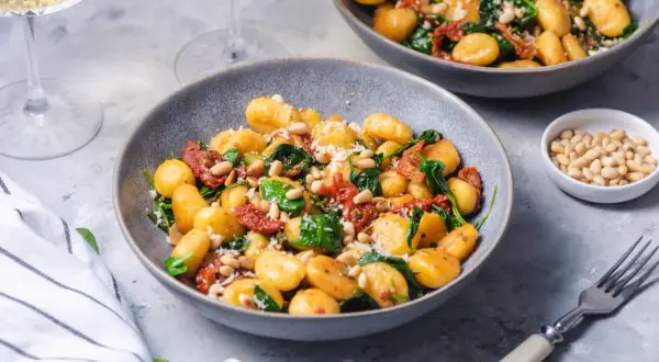 Pyszne gnocchi ze szpinakiem, suszonymi pomidorami i orzeszkami pinii, posypane parmezanem.