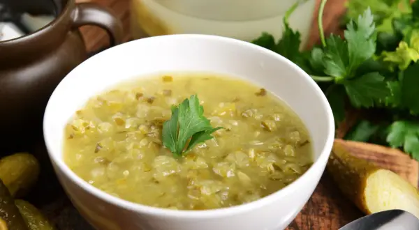 Zupa ogórkowa przepis babci. Aromatyczna, domowa zupa z kiszonych ogórków, podana w białej miseczce z gałązką natki pietruszki. Obok leżą ogórki kiszone.