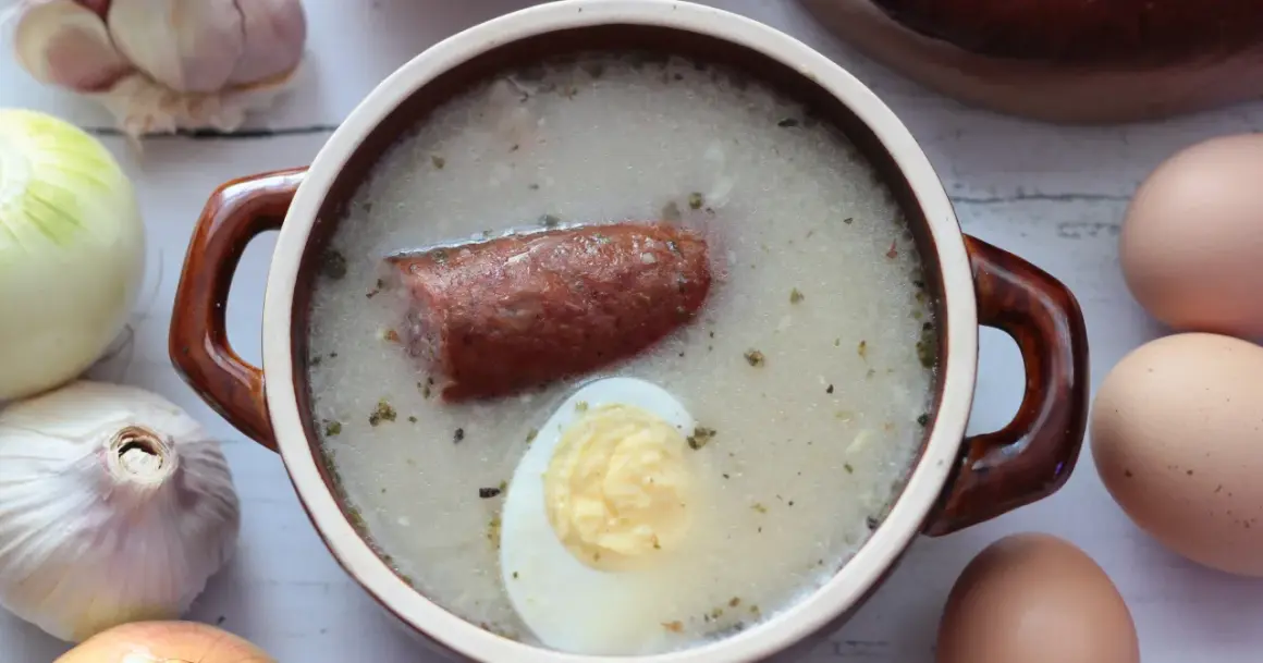 Aromaticzny żurek z kiełbasą i jajkiem, gotowy do podania. W tle czosnek, cebula i jajka, składniki idealnego zakwasu na żurek.