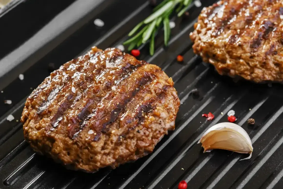 Soczyste mięso na burgery grilluje się na patelni, obok gałązka rozmarynu i ząbek czosnku.
